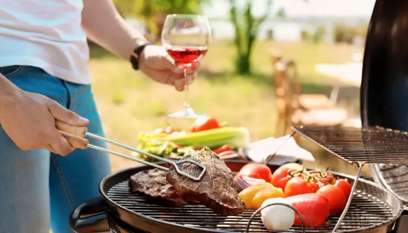 Un homme savoure un verre de vin rouge tout en préparant un barbecue avec des steaks et des légumes.