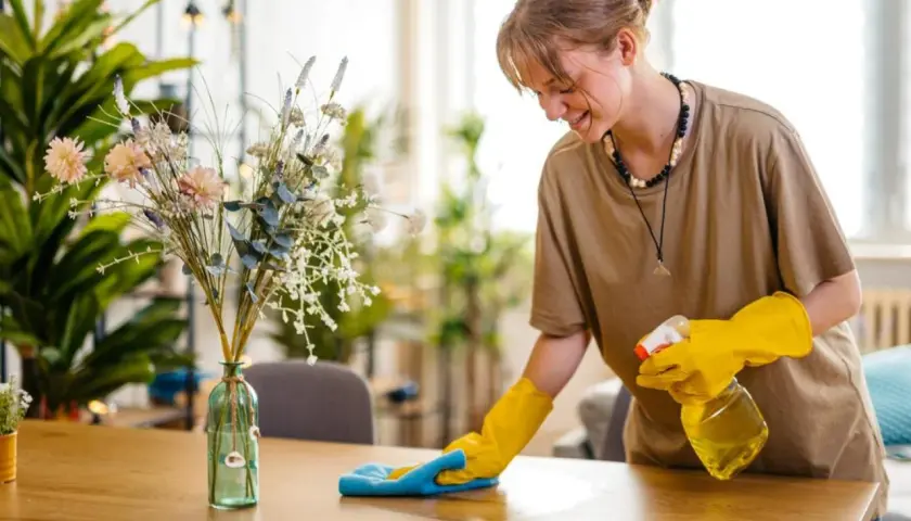 Joven sonríe mientras aprende como limpiar el polvo de los muebles con spray y paño azul.