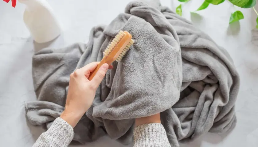 A person is brushing a grey throw blanket, showing how to wash it. A spray bottle is nearby.