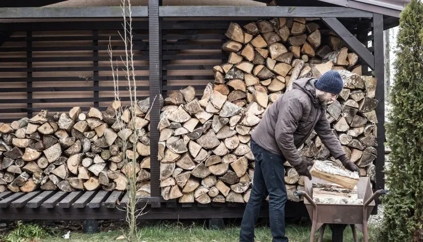 Mann mit blauer Mütze lagert Holz in einer Schubkarre vor einem Holzschuppen.