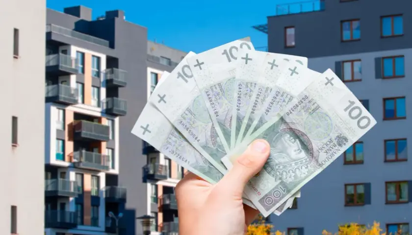 Dłoń trzyma polskie banknoty na tle nowoczesnych budynków mieszkalnych. Jak uniknąć podatku od sprzedaży nieruchomości przed upływem 5 lat?