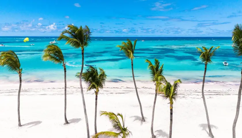 Traumhafter **Urlaub im November** an einem karibischen Strand mit Palmen, weißem Sand und türkisfarbenem Meer. Boote und Parasailing im Hintergrund.