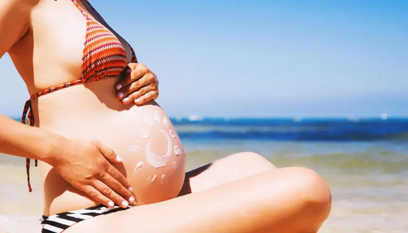 Brzuszek w ciąży na plaży, posmarowany kremem z filtrem w kształcie słońca. Bezpieczne opalanie w ciąży to podstawa.