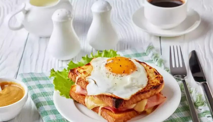 Un délicieux croque monsieur garni d'un œuf au plat, servi avec une tasse de café et une sauce crémeuse.