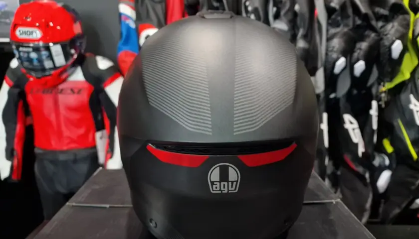 Matowy kask AGV z subtelnym wzorem, gotowy na przygodę. W tle czerwono-czarny kombinezon motocyklowy.