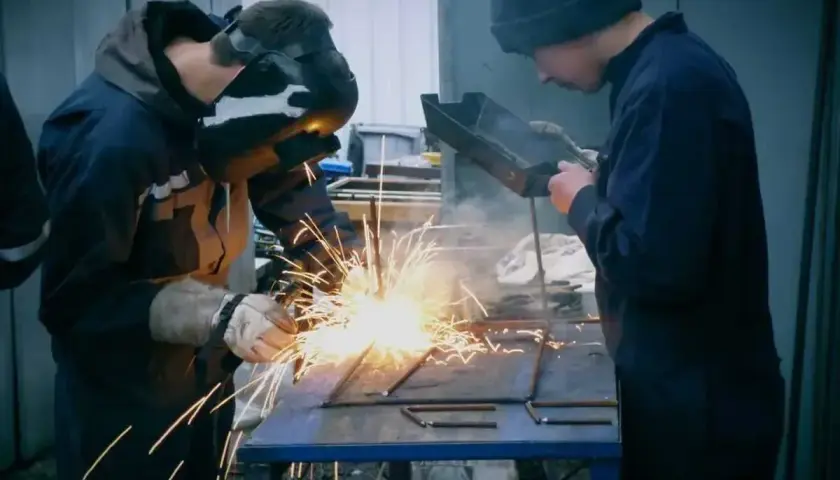 Dwóch spawaczy pracuje przy metalowej konstrukcji. Jeden spawa elektrodą, tworząc snop iskier. Drugi obserwuje, trzymając narzędzie.