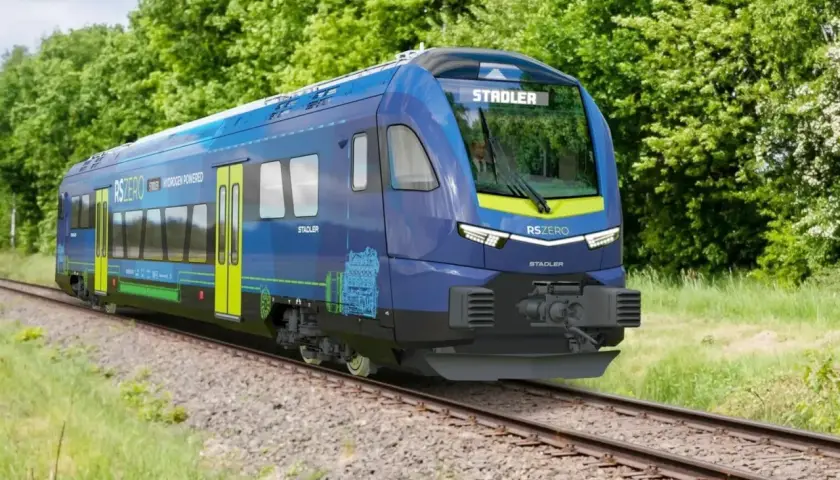 Niebieski pociąg Stadler RSZERO na wodór jedzie po torach wśród zieleni.