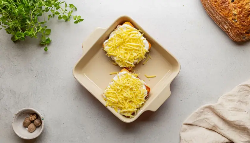 Dwa tosty z szynką i serem, przygotowane do zapieczenia, jak w klasycznym croque madame. Obok gałka muszkatołowa i świeża zielenina.