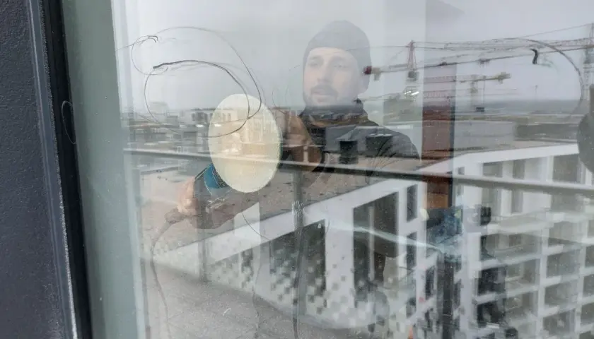 Mężczyzna w czapce wykonuje polerowanie szkła za pomocą maszyny. W odbiciu widać budynki i dźwigi.