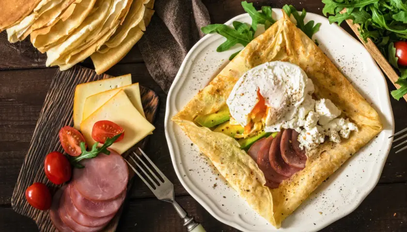 Złożone crepes z jajkiem w koszulce, awokado, szynką i serem. Obok stos naleśników, pomidorki i rukola.