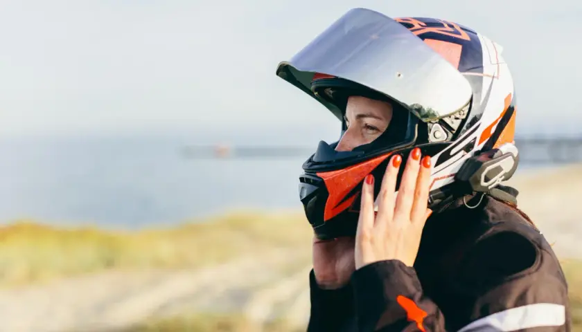 Donna con casco da moto colorato, con mani che aggiustano la visiera per evitare che si appanni. Sfondo sfocato di mare e sabbia.