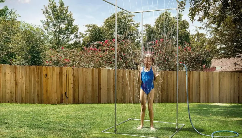 Ein Mädchen im blauen Badeanzug genießt eine erfrischende Dusche unter einer selbstgebauten Gartendusche im sonnigen Garten.