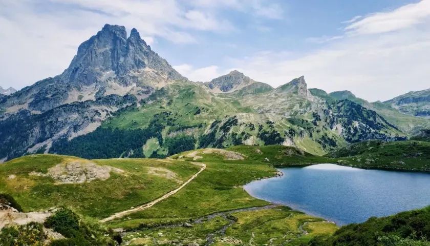 Majestuoso pico en el valle de Ossau, con un lago azul y senderos serpenteantes entre verdes colinas.