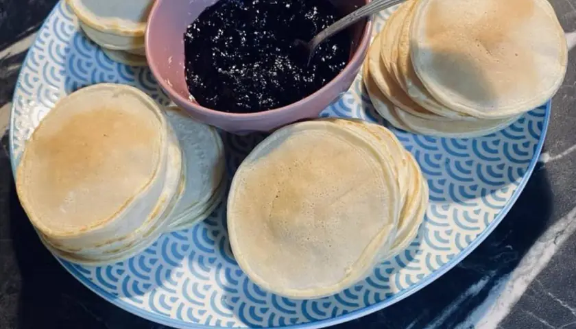 Une assiette bleue et blanche présente des piles de poffertjes dorés, accompagnés d'un bol de confiture de myrtilles. Parfait pour une recette de poffertjes !