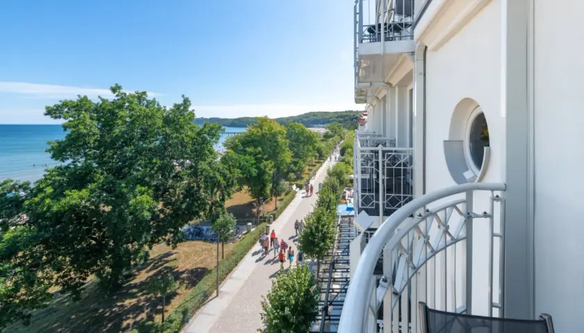 Blick von einem Balkon auf die belebte Binz Promenade mit Bäumen, Meer und Menschen, die spazieren gehen.