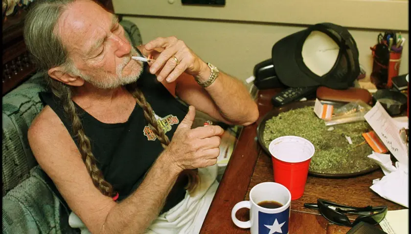 Willie Nelson, icona delle canzoni country famose, si accende una sigaretta con una tazza di caffè accanto.