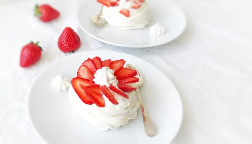 Deux pavlovas à la fraise, légers et aériens, ornés de fruits rouges frais et de crème fouettée, sur des assiettes blanches.