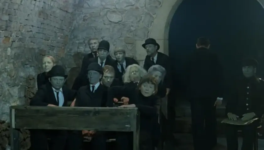 Postacie z "Umarłej klasy" Tadeusza Kantora, w tym uczniowie i nauczyciele, siedzą w ponurej scenerii.