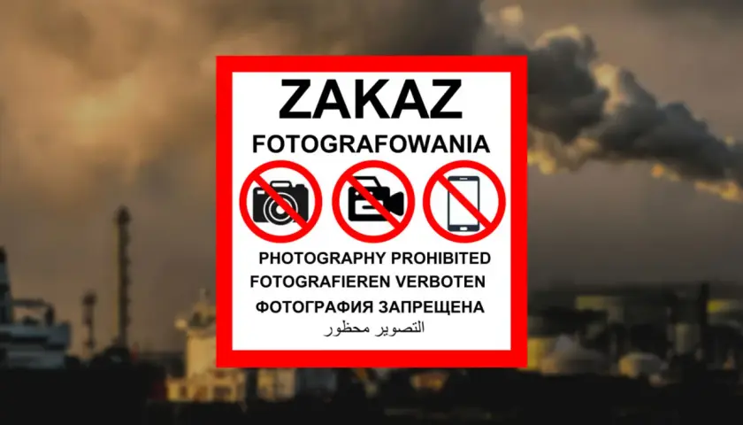 Tablica z napisem "ZAKAZ FOTOGRAFOWANIA" i symbolami przekreślonych aparatów, kamer i telefonów komórkowych. W tle widać przemysłowy krajobraz z kominami fabrycznymi i statkami. To znak informujący, czego nie można fotografować w Polsce.