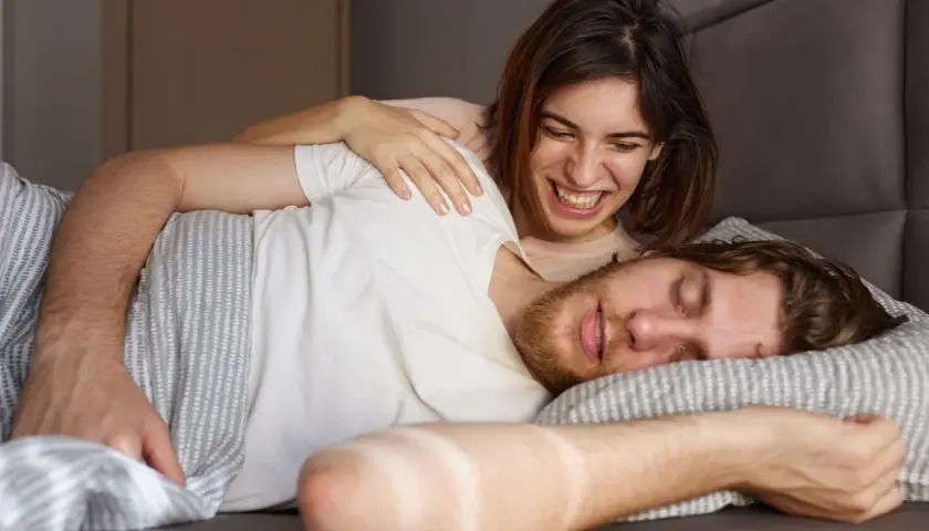 Ein Mann schläft, während eine Frau ihn lachend kitzelt. Vielleicht redet er im Schlaf und sie amüsiert sich.