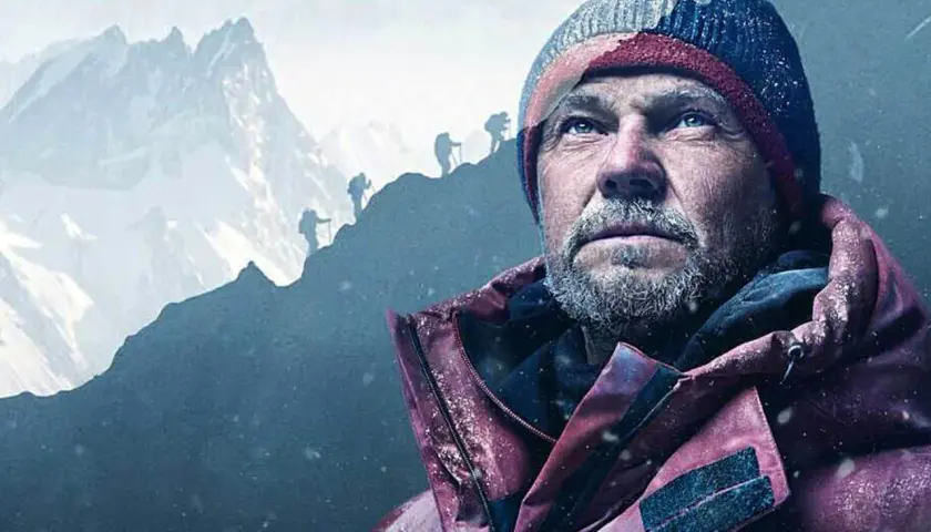 Zmęczony alpinista w czapce i kurtce patrzy w górę. W tle widać innych wspinaczy na zboczu podczas wyprawy na Broad Peak.