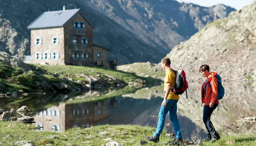 Zwei Wanderer auf einer Hufeisentour im Sarntal, vorbei an einer Berghütte und einem Bergsee.