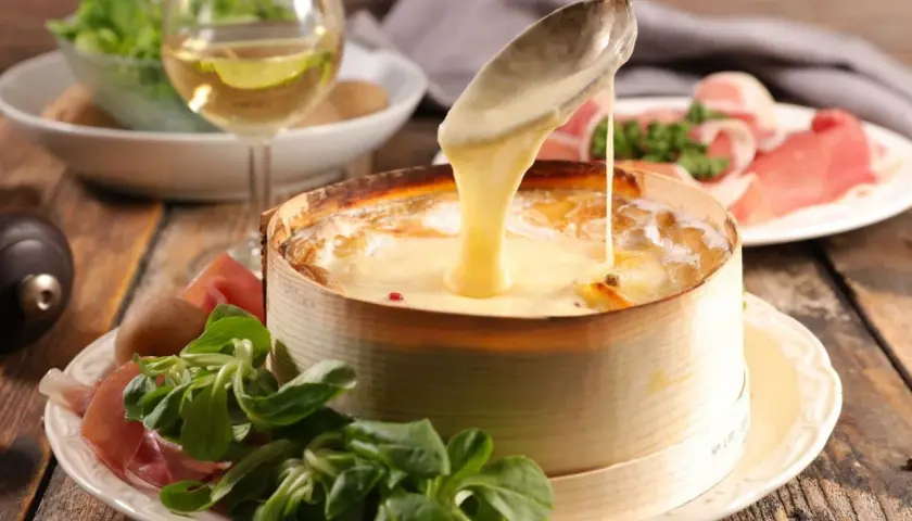Un Mont d'Or fondant est servi avec une cuillère, accompagné de charcuterie, de pommes de terre et d'une salade verte. Un verre de vin blanc complète ce plat gourmand.