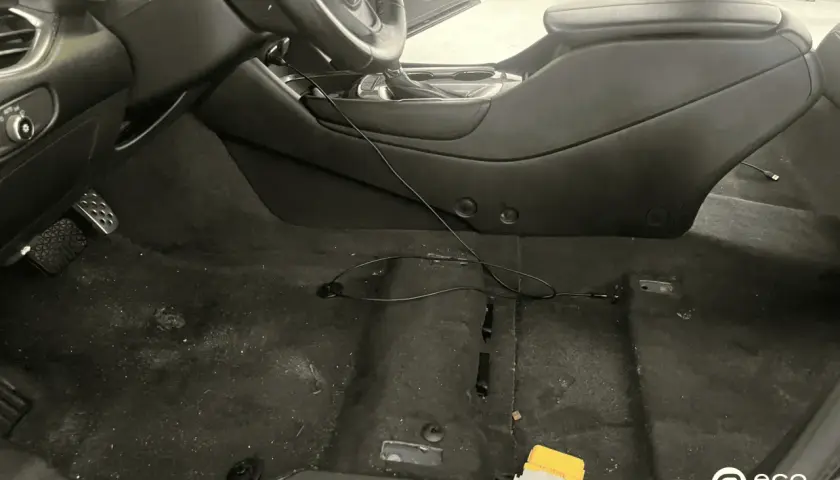 Interior de coche con moqueta oscura y algo de suciedad. Se ve el volante, consola central y pedales. Hay cables y un pequeño objeto amarillo.