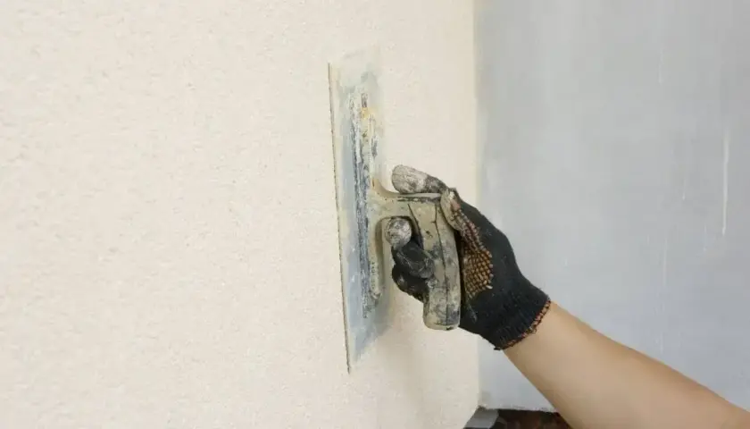 Ręka w rękawicy nakłada tynk kornik na ścianę, tworząc charakterystyczną fakturę.