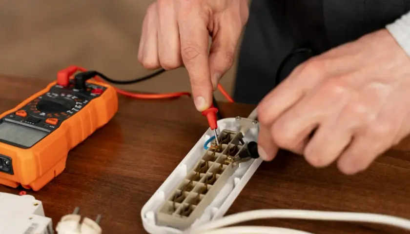 Ręce mierzą opór elektryczny w przedłużaczu za pomocą pomarańczowego multimetru.