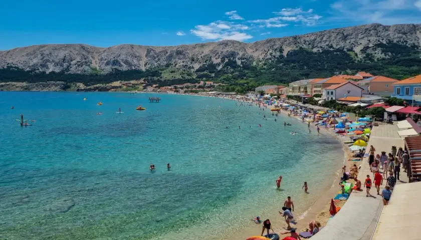 Słoneczna plaża na wyspie Krk, pełna ludzi kąpiących się i opalających. W tle góry i zabudowania. To idealne miejsce na wakacyjne atrakcje.