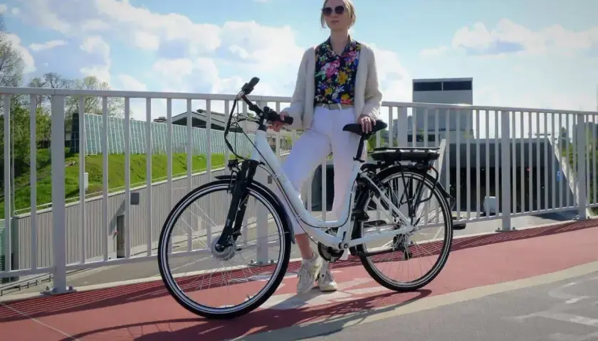 Kobieta w białych spodniach i kolorowej koszuli stoi obok białego roweru elektrycznego. Czy na rower elektryczny trzeba mieć prawo jazdy?