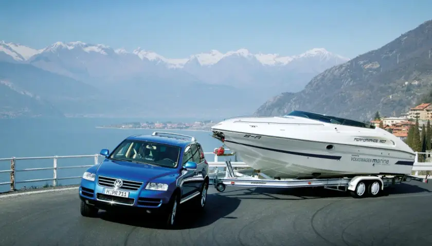 Un coche azul remolca un barco deportivo blanco por una carretera junto a un lago y montañas nevadas.