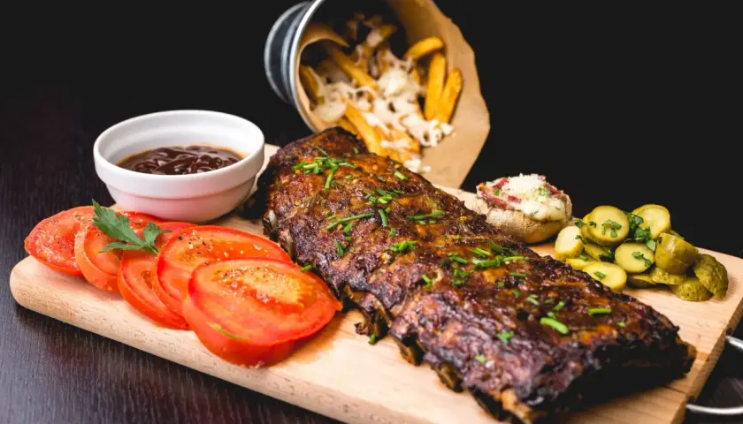 Un plat de côtes de porc au barbecue, garni de ciboulette, servi avec des tomates tranchées, des cornichons, une sauce barbecue et des frites au fromage.