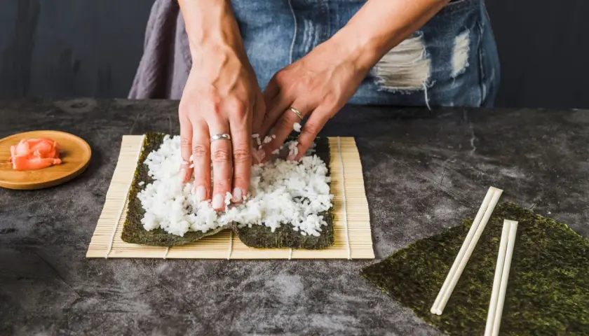 Dłonie rozkładają ryż na nori na macie bambusowej do sushi.