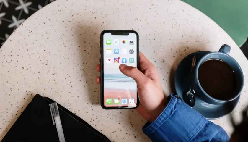 Main d'une personne tenant un smartphone affichant des icônes d'applications, comme un exemple d'application web. Une tasse de café est à côté.