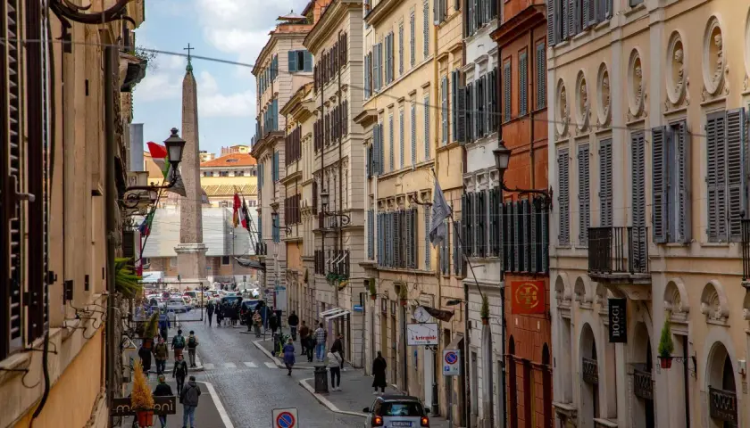 Una strada romana con palazzi storici, un obelisco e gente che passeggia. Si intuisce quanto è lunga Via del Corso, con la sua prospettiva che si perde all'orizzonte.