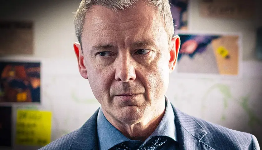 En man i kostym, med grånande hår, blickar fundersamt framåt. Han är en del av grace serie.