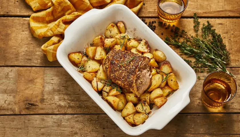 Selle d'agneau rôtie avec pommes de terre dorées et herbes aromatiques dans un plat blanc, prête à être dégustée.