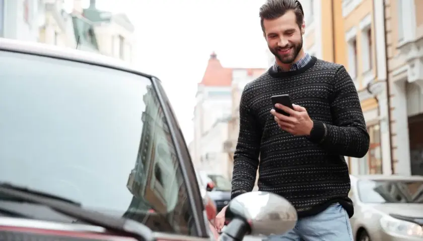 Uśmiechnięty mężczyzna z telefonem w ręku stoi obok czerwonego samochodu na ulicy.