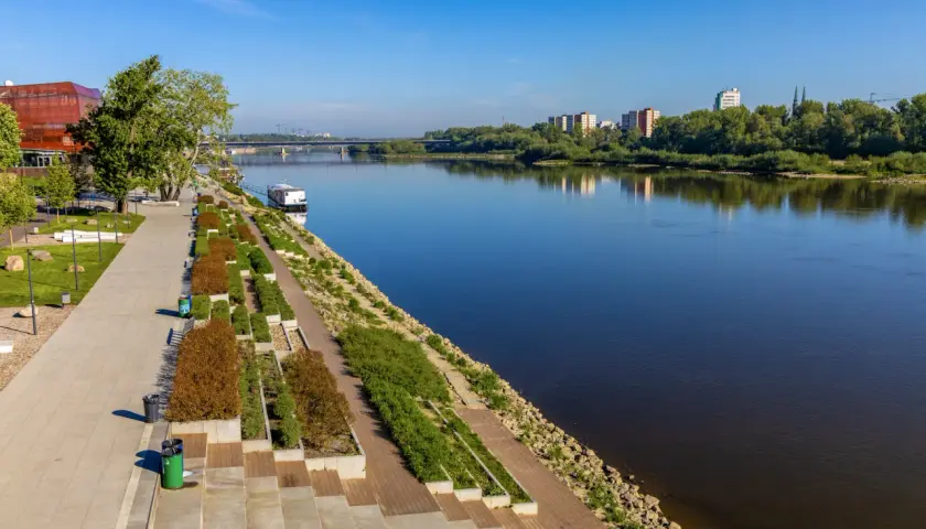Nowoczesny boulevard nad rzeką z zielenią, nowoczesnym budynkiem i mostem w oddali.