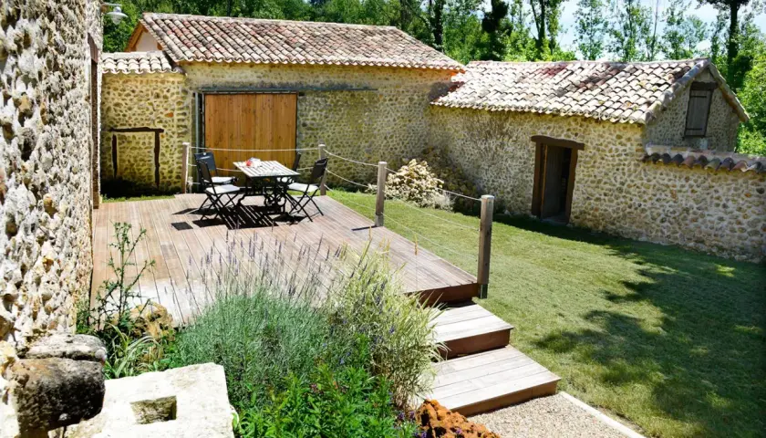Aménagement longère : terrasse en bois avec mobilier, jardin verdoyant et murs en pierre.