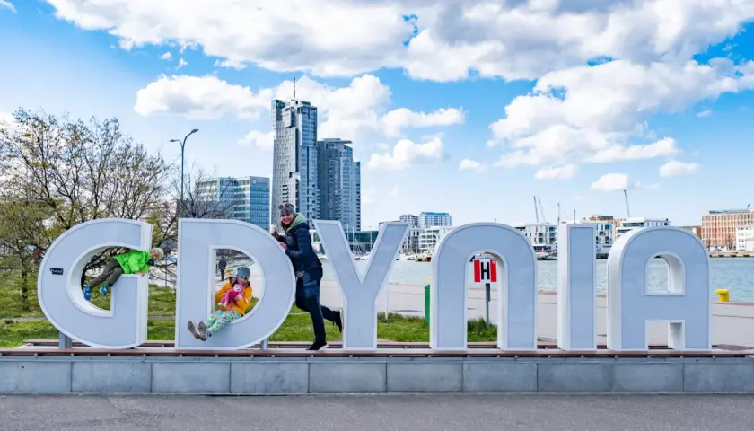Dzieci bawią się na literach napisu "GDYNIA" na tle miejskiego krajobrazu.