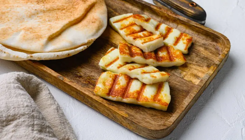 Des morceaux de halloumi grillé, marqués par les rayures du grill, reposent sur un plateau en bois à côté d'un pain pita moelleux.