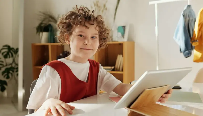 Chłopiec z kręconymi włosami uśmiecha się, trzymając tablet. Czy to wady i zalety szkoły waldorfskiej?