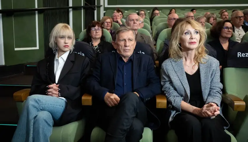 Małgorzata Englert, w wieku, siedzi w pierwszym rzędzie obok innych osób, wpatrując się w dal.