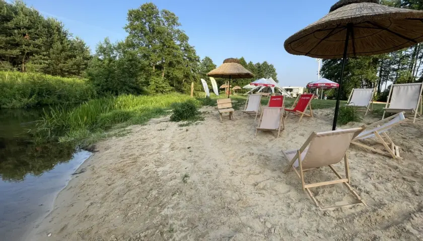 Plaża nad rzeką z leżakami i parasolami. W tle drzewa i niebo. Idealne miejsce na relaks, jak na konopnicy.