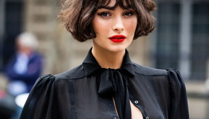 Mujer con un elegante corte de pelo **french bob**, labios rojos vibrantes y una blusa negra transparente con lazada.