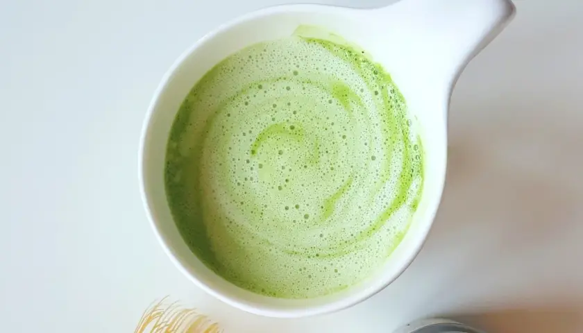 Zielony, spieniony napój matcha w białej miseczce, idealny jako alternatywa co zamiast kawy. Obok bambusowa miotełka i proszek matcha.