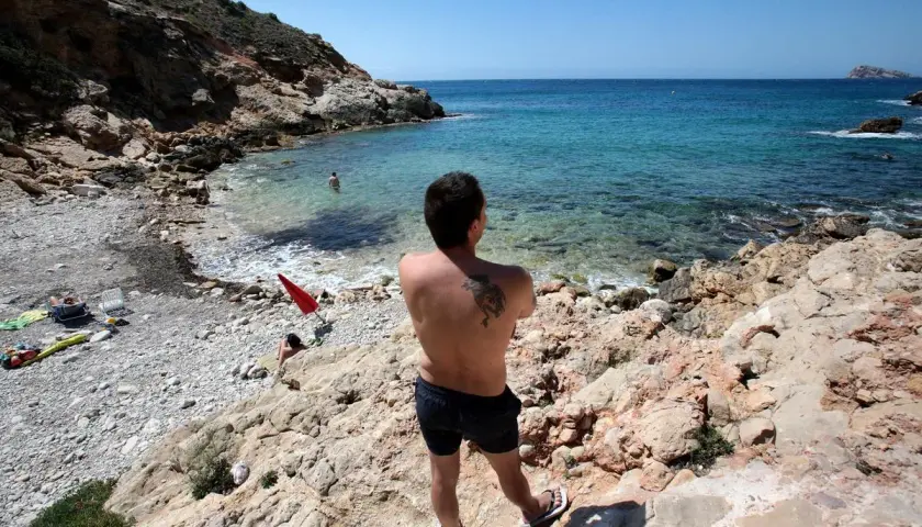 Hombre con tatuaje en la espalda disfruta de la vista en la playa nudista Benidorm. Aguas cristalinas y rocas caracterizan este rincón.